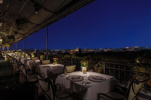 Un restaurant où les tables sont dressées pour le dîner, avec une vue sur le paysage urbain illuminé la nuit.