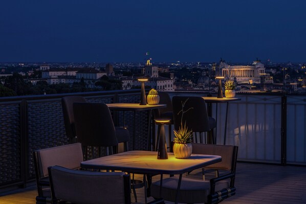 Una terrazza sul tetto con tavoli e sedie, una pianta in vaso e vista sulla città al crepuscolo.