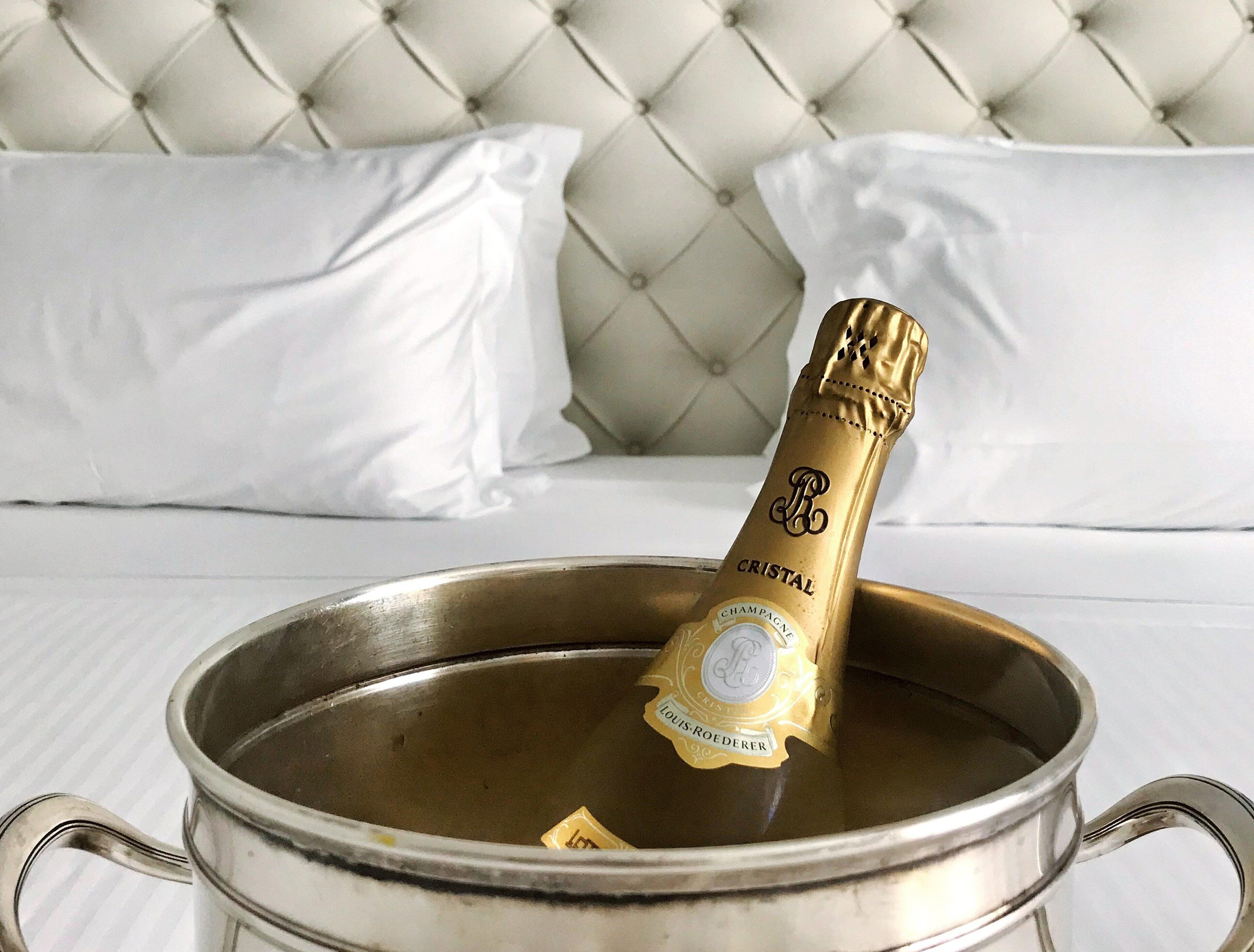A champagne bottle in a metal bucket on a bed with white pillows and a tufted headboard.
