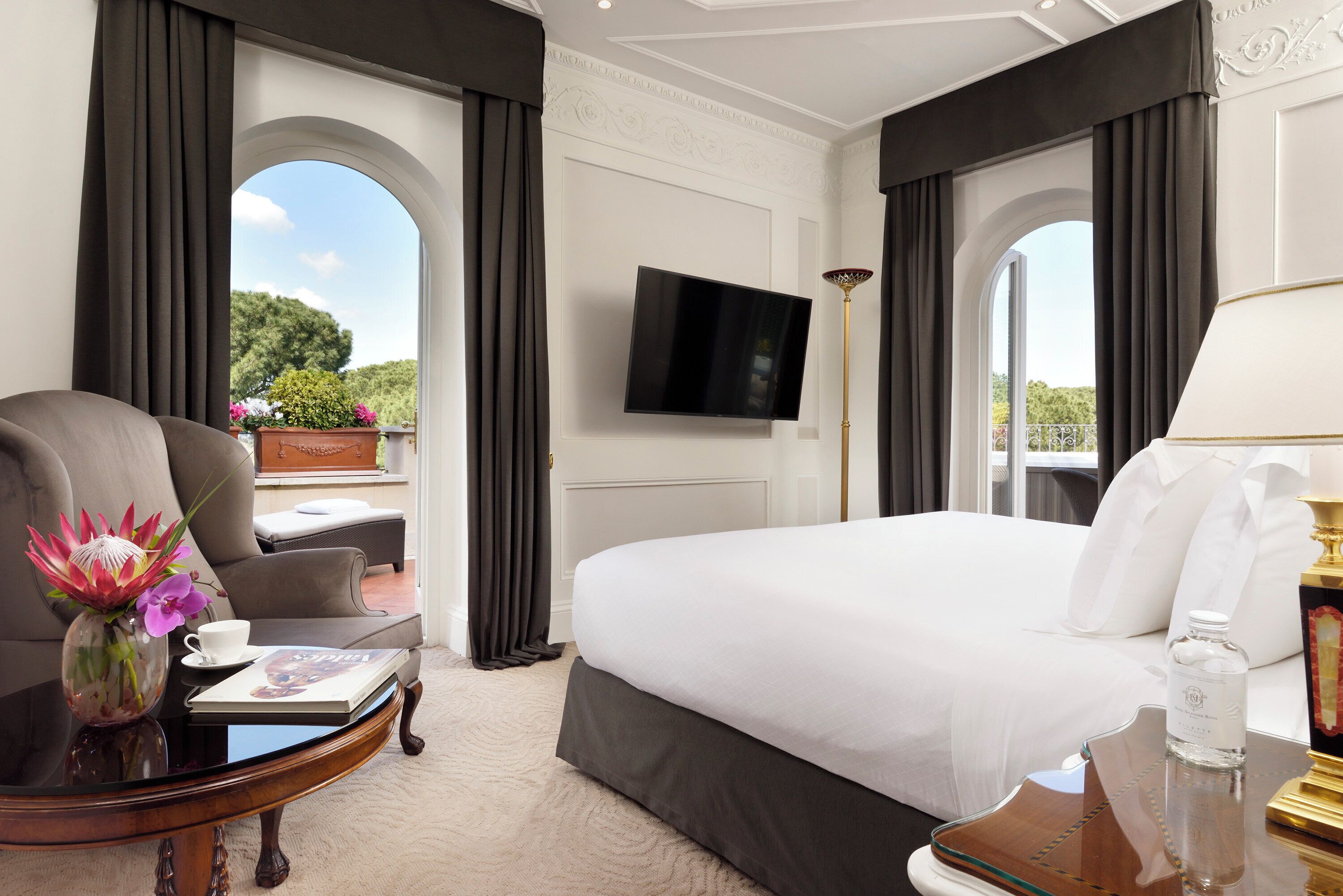 A hotel room with a large bed, a flat-screen TV, a coffee table with a vase of flowers, and a view of the outdoors.
