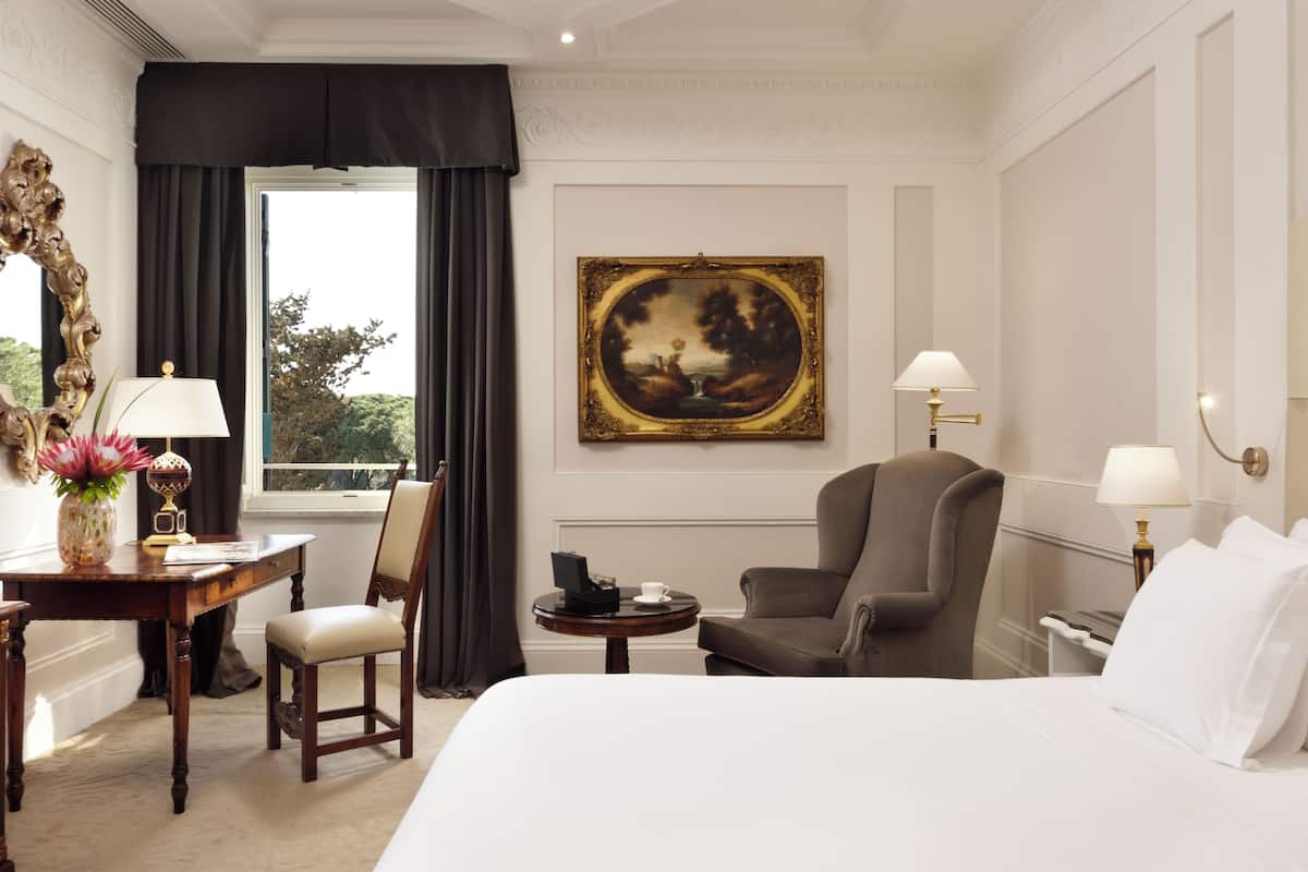 Ein Hotelzimmer mit einem großen Bett, einem Schreibtisch inklusive Lampe, einem Sessel, einem kleinen Tisch ebenfalls mit Lampe und einem Bild an der Wand.