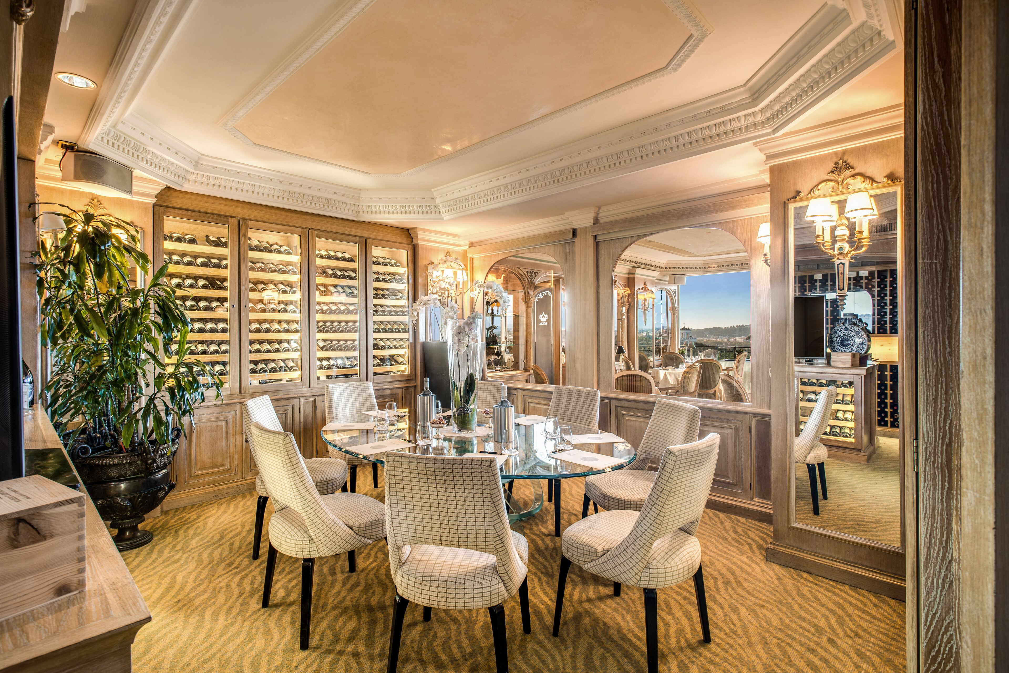 A luxurious dining area with a glass table surrounded by upholstered chairs, a potted plant, and a wine rack in the background.