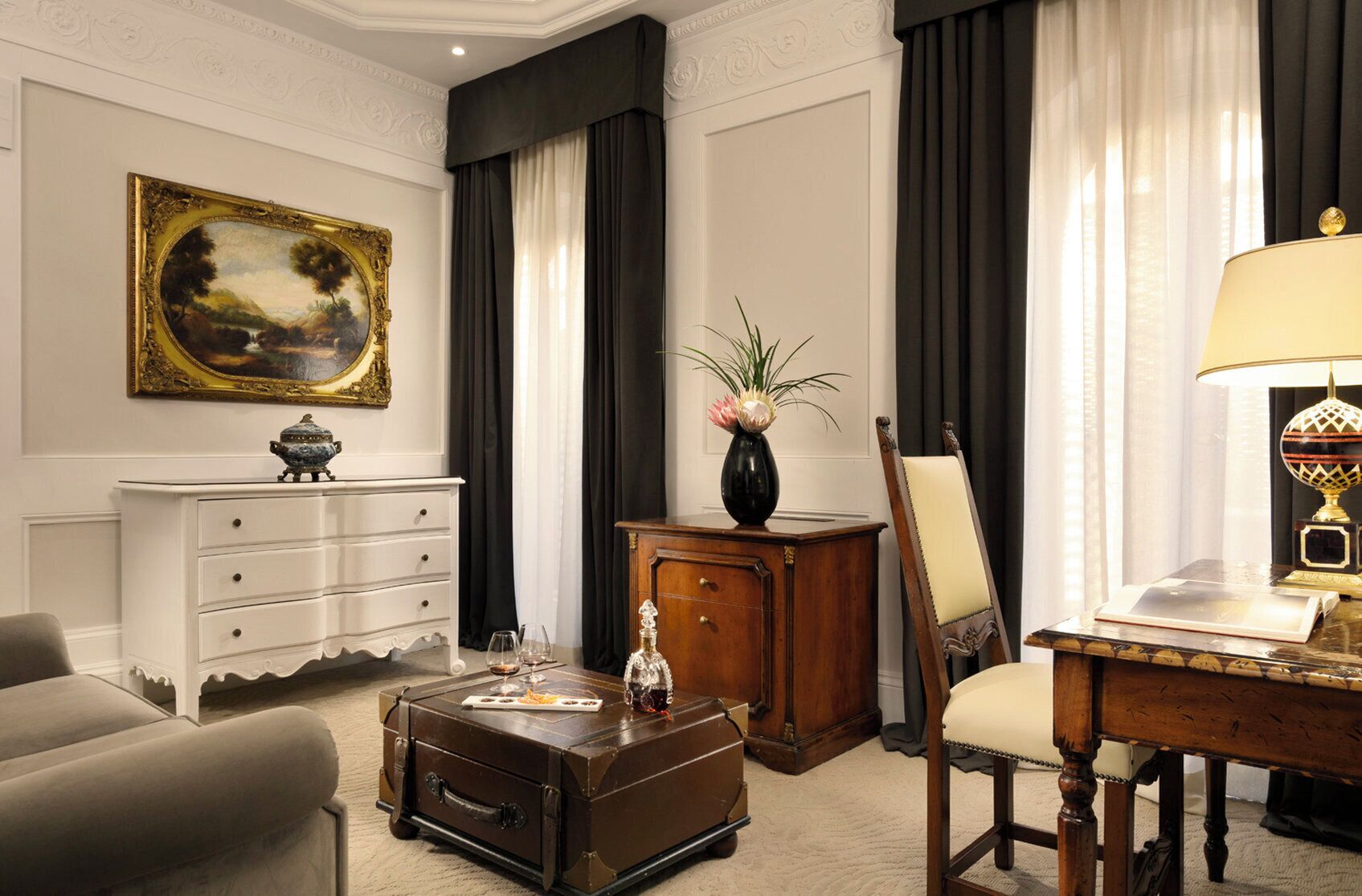 A room with a vintage-style desk, a chair, a small table with a lamp, a cabinet, a painting on the wall, and a vase with flowers.