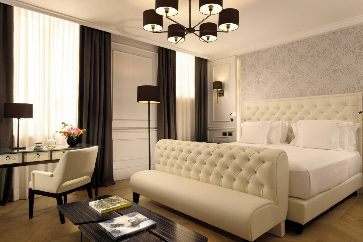 Ein Hotelzimmer mit einem großen Bett, einem Schreibtisch mit Stuhl, einem Nachttisch, einer Lampe und Blick ins Freie durch das Fenster.