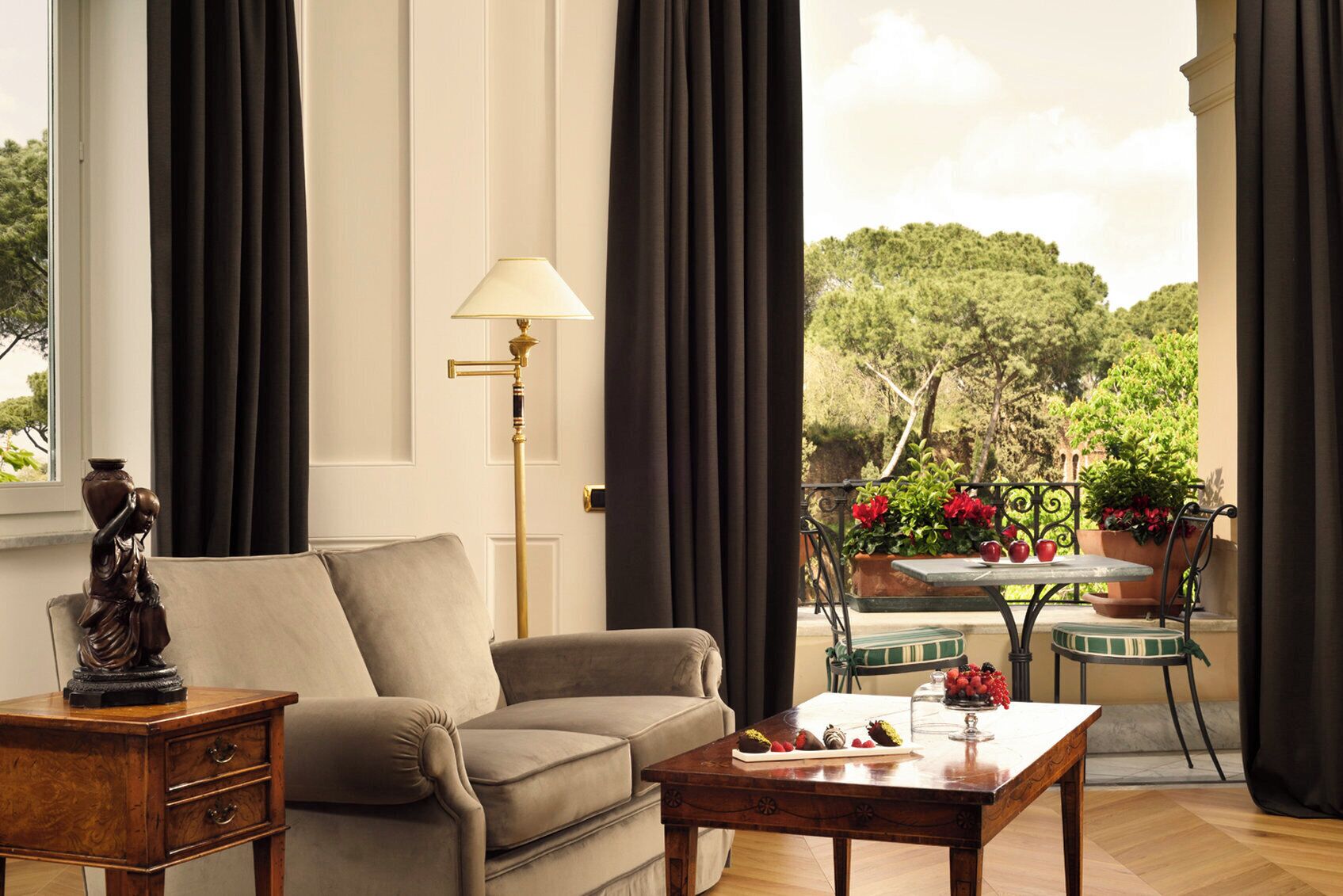 A living room with a sofa, a lamp, a statue, and a view of a balcony with potted plants and a table.