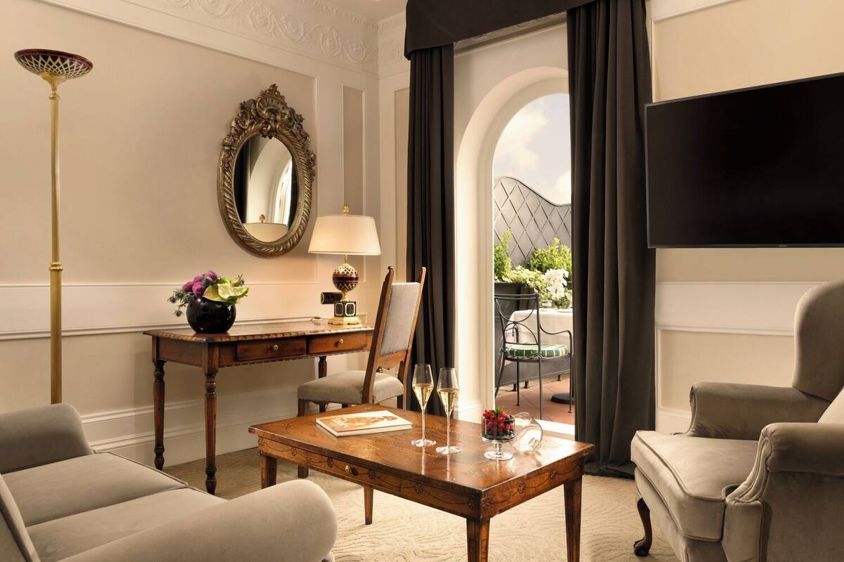 Ein Hotelzimmer mit einem Holztisch, zwei Sesseln, einer Couch, einem großen Spiegel, einem Flachbildfernseher und Blick auf einen Garten.