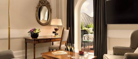 A hotel room with a wooden desk, two armchairs, a sofa, a large mirror, a flat-screen TV, and a view of an outdoor garden.