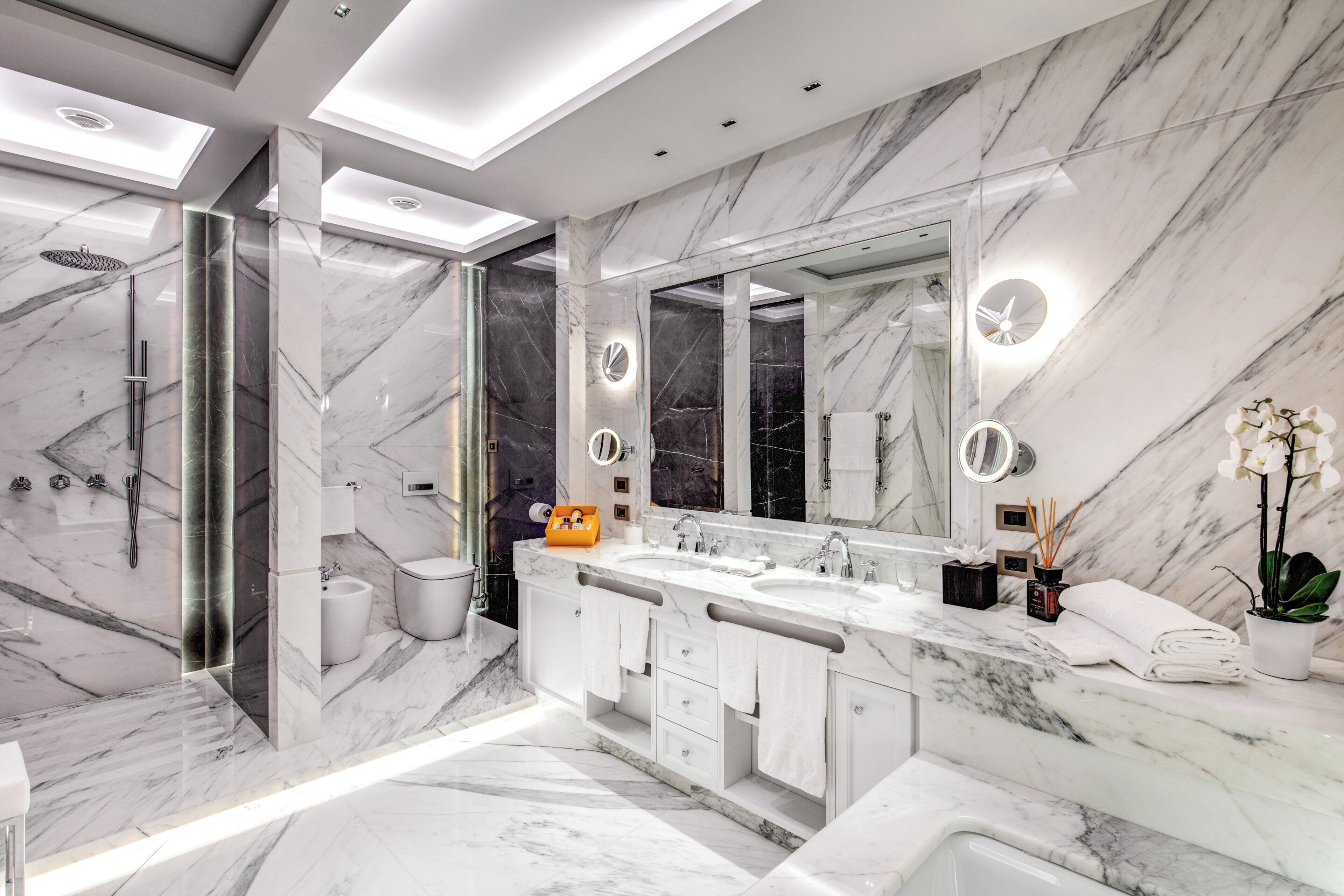 A modern bathroom with a large mirror, two sinks, a shower, and marble walls.