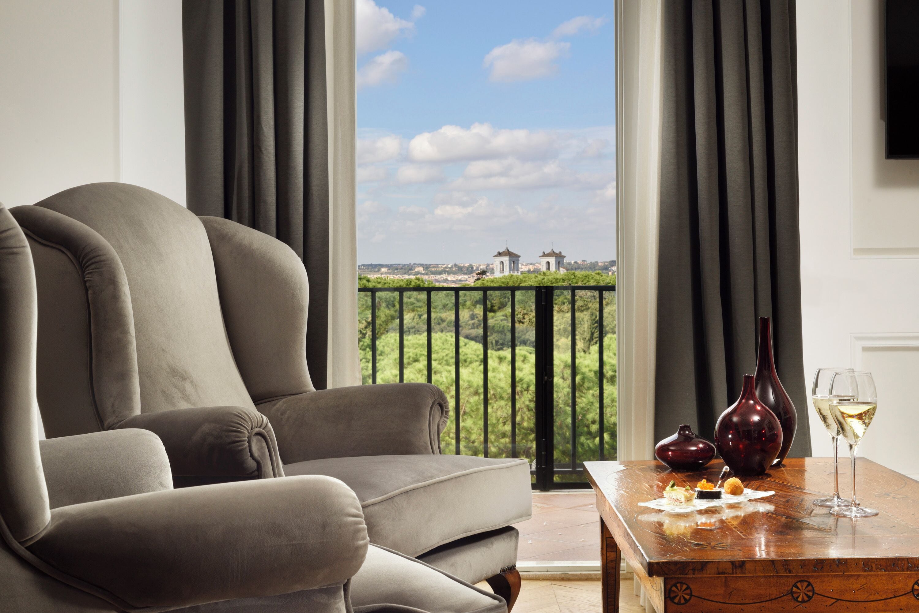 A modern living room with a grey sofa, a wooden table with a vase and wine glass, and a balcony with a view of greenery and buildings.