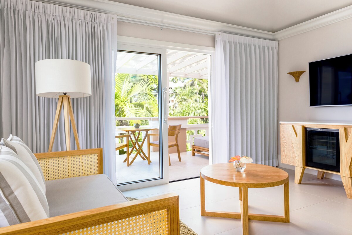 Un salon moderne comprenant un canapé, une lampe sur pied, une table basse ronde, un téléviseur et une porte-fenêtre coulissante donnant sur une terrasse extérieure avec une table et des chaises.