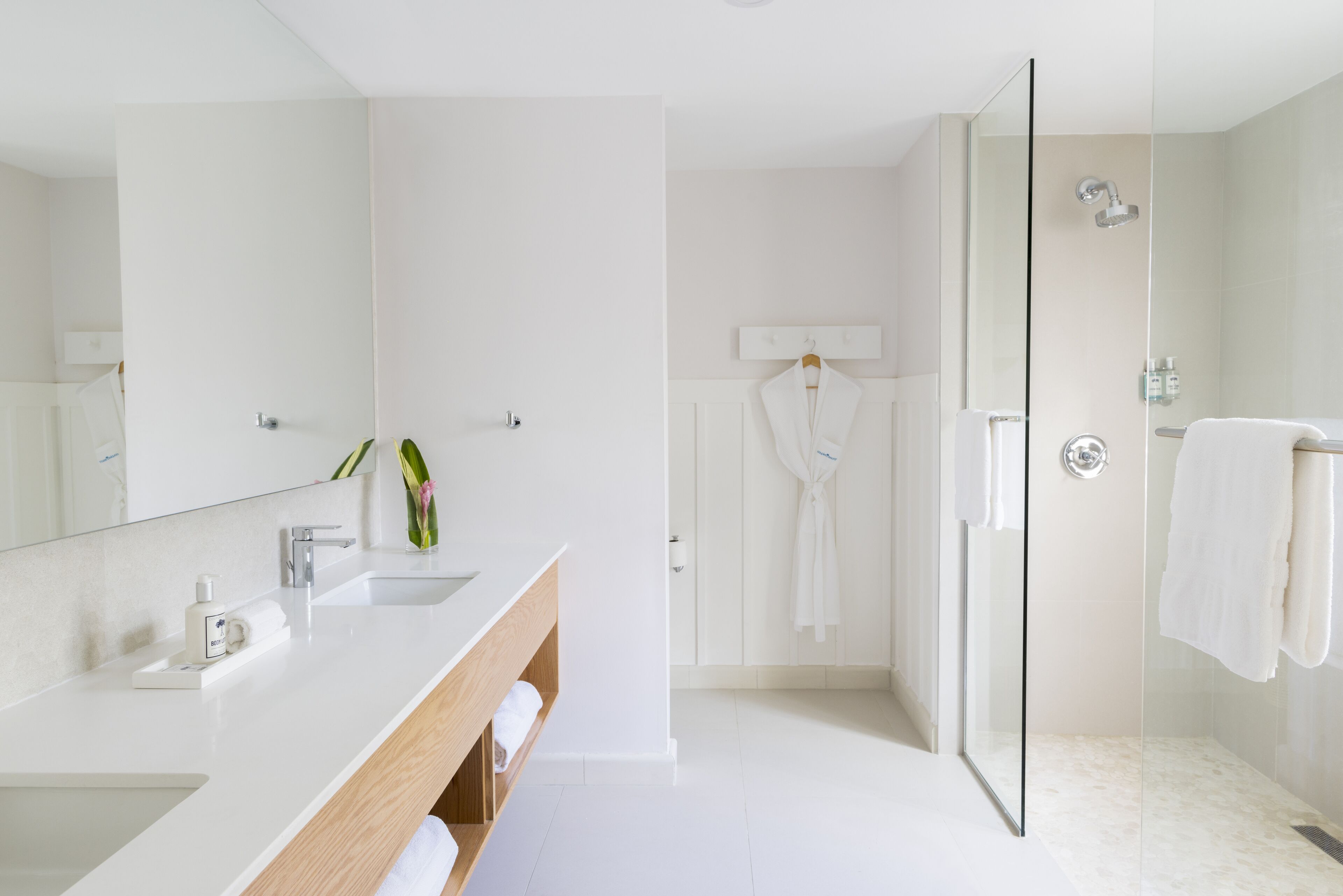A modern bathroom with a large mirror, a white sink, a glass shower enclosure, and a robe hanging on a hook.
