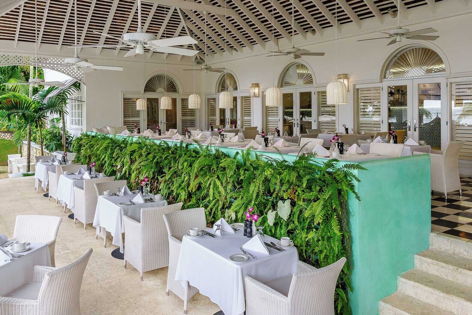 Un amplio comedor interior con mesas y sillas blancas, un separador verde y ventiladores de techo.