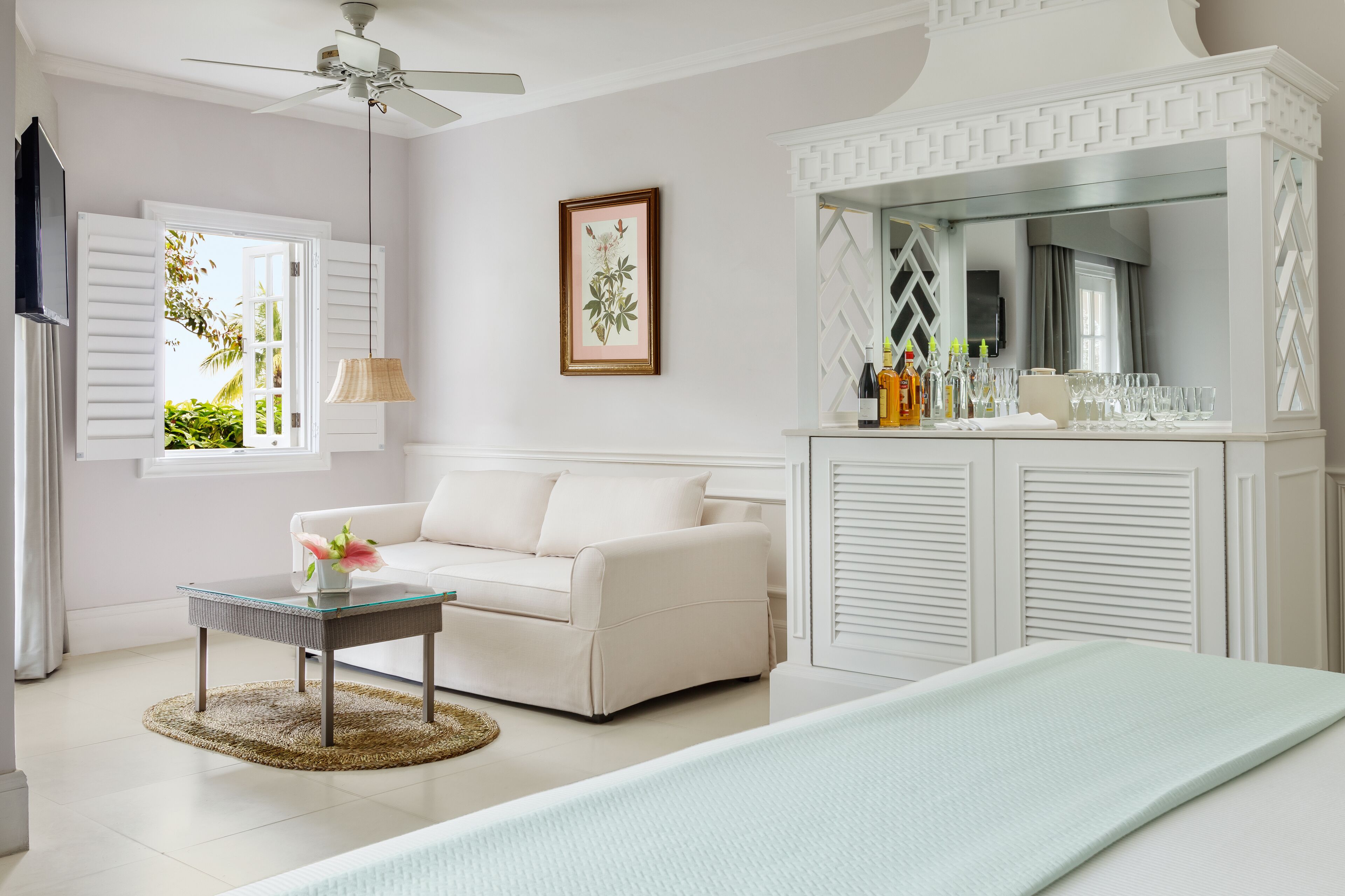 A room with a white sofa, a glass coffee table, a bed with a light-colored bedspread, a ceiling fan, a framed botanical print, and a minibar with bottles and glasses.