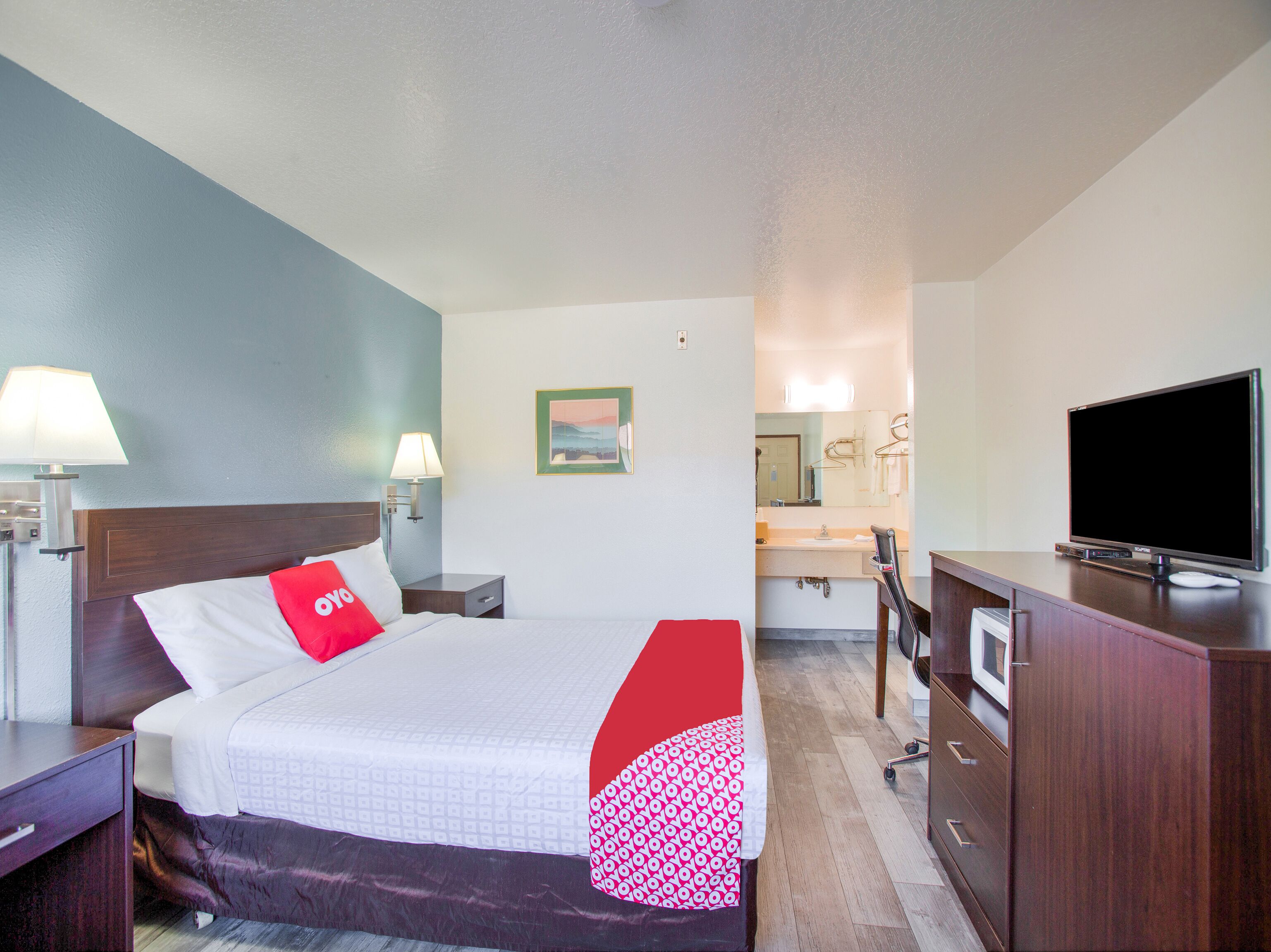 A hotel room with a bed, bedside lamps, a TV, a desk, and a bathroom visible through an open door.