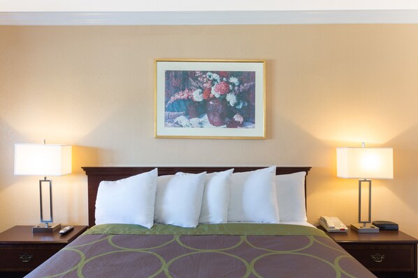 Habitación de hotel con cama, dos mesitas de noche, lámpara, control remoto y un cuadro de flores en la pared.