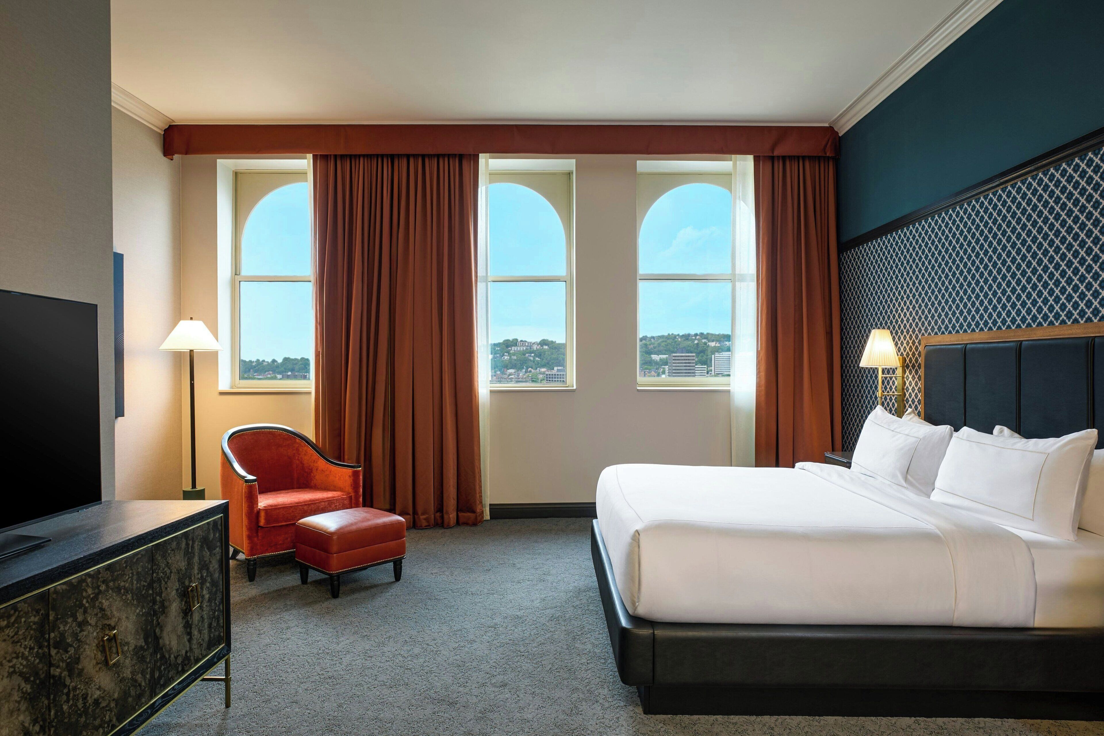 Une chambre d’hôtel avec un lit, une chaise, une télévision et des fenêtres avec des rideaux.