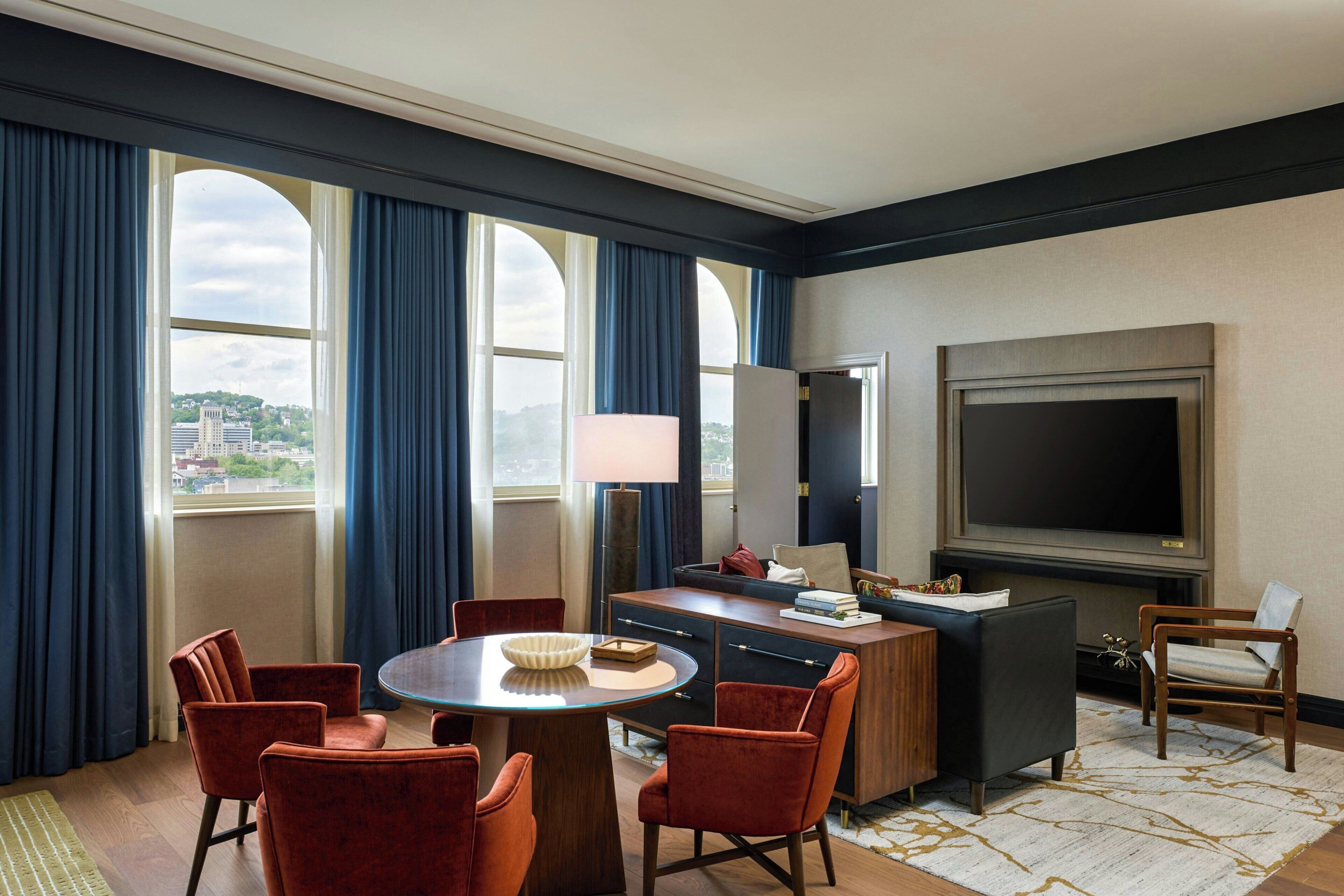 A hotel room with a large window, a flat-screen TV, a dining table with chairs, and a sofa.