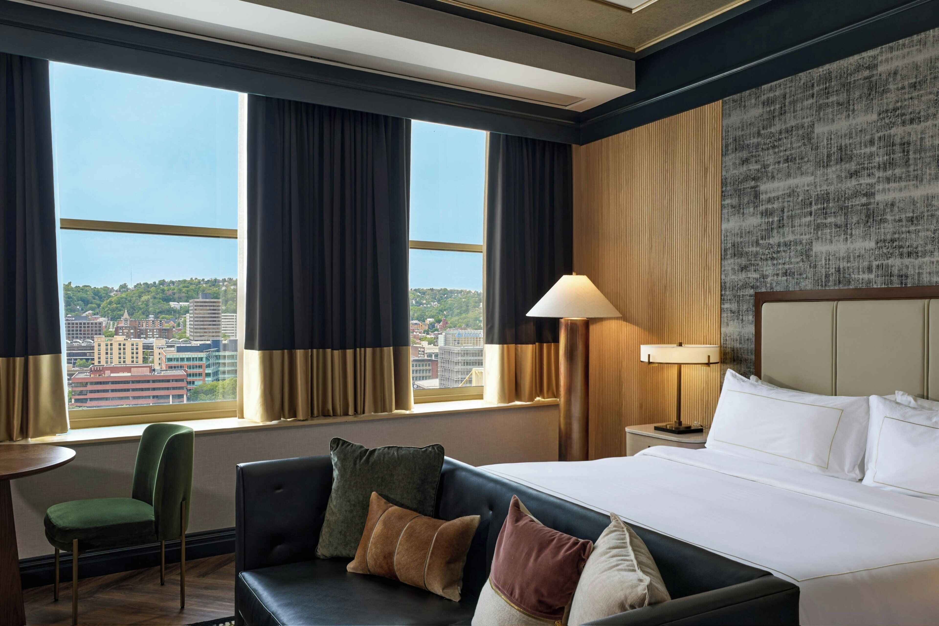 A hotel room with a bed, a green chair, a sofa, and a view of the cityscape through large windows.
