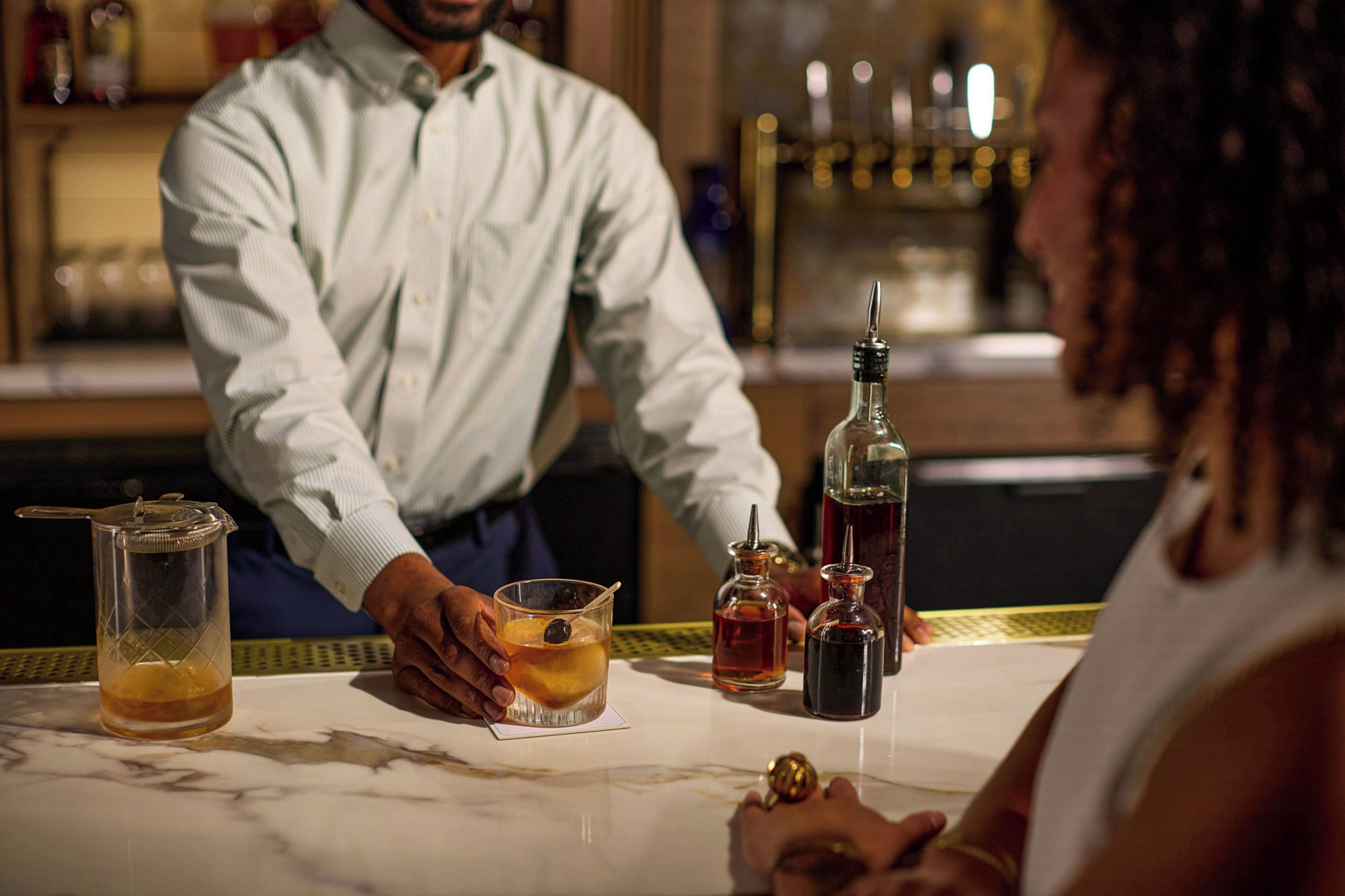 A bar setting with a bartender and a customer. There are various bottles and a glass on the counter.