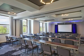 Une salle de conférence avec des rangées de tables et de chaises, de grandes fenêtres et un écran de projection.