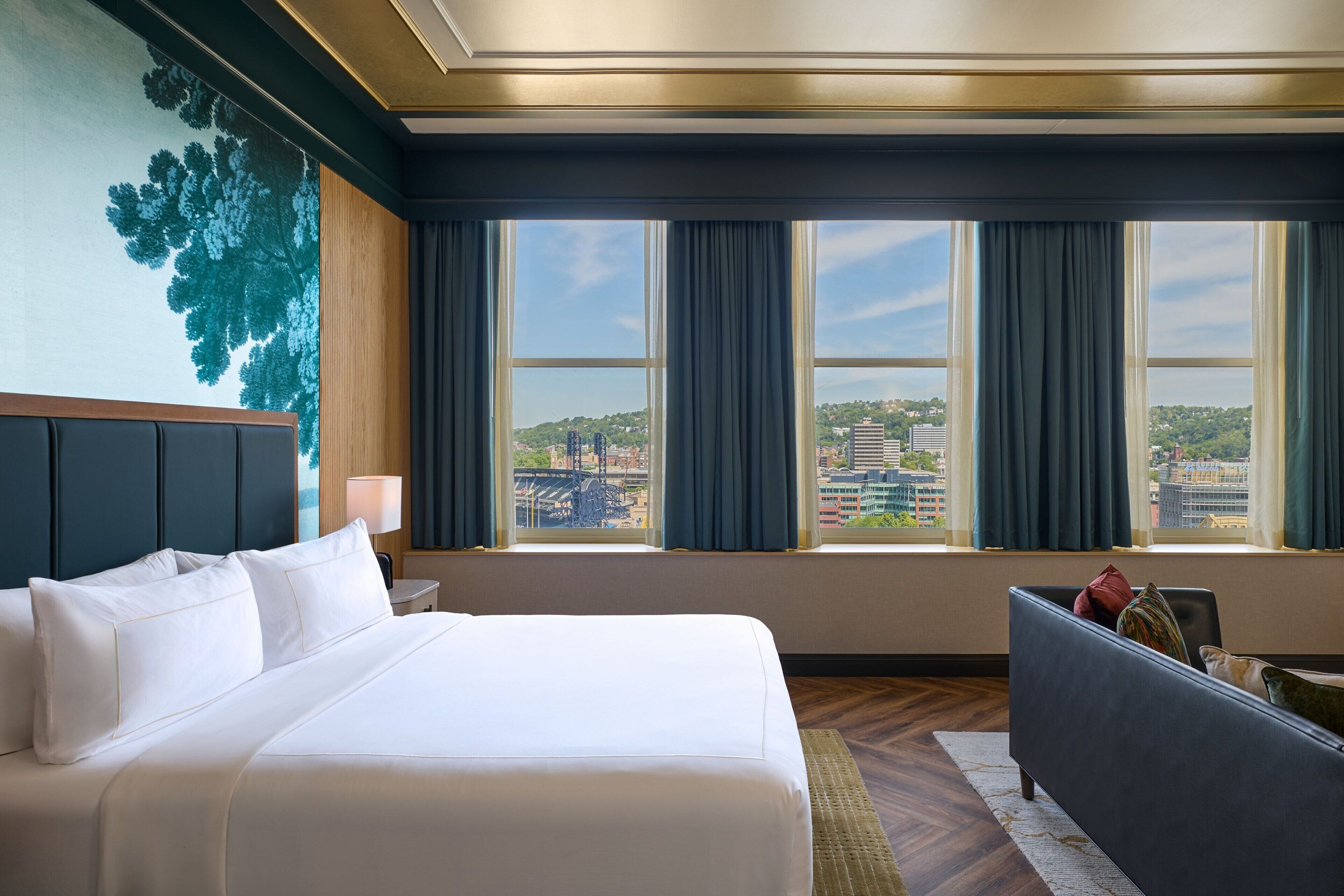 A hotel room with a large bed, a desk, a chair, and a view of the cityscape through the windows.