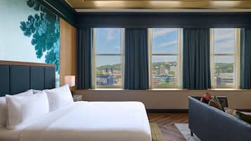 A hotel room with a large bed, a desk, a chair, and a view of the cityscape through the windows.