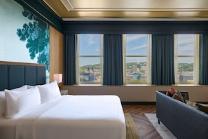 A hotel room with a large bed, a desk, a chair, and a view of the cityscape through the windows. - The Atterbury Hotel, Autograph Collection (Pittsburgh)