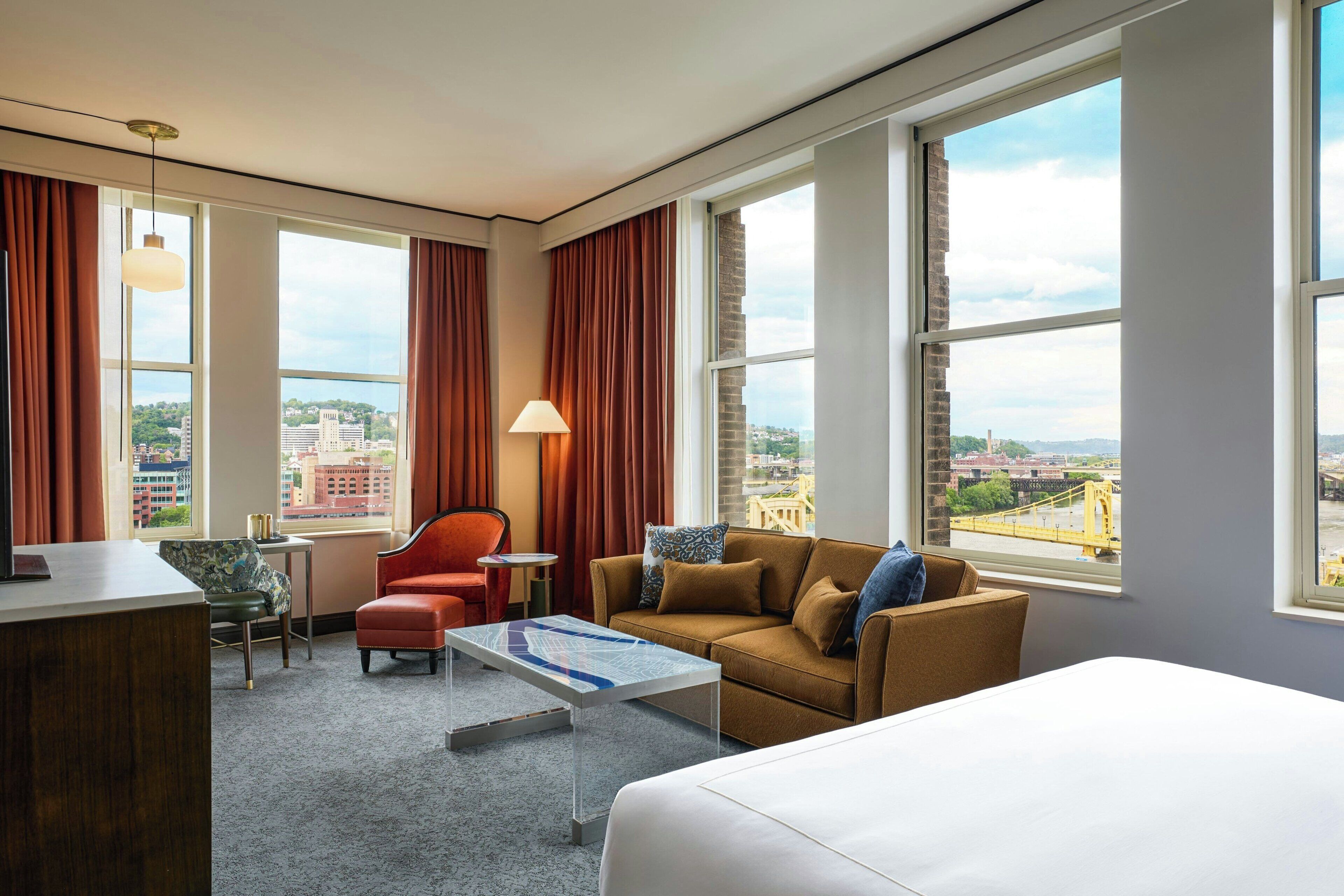 Quarto de hotel com cama, sofá, poltrona, mesa pequena e abajur. Janelas amplas com cortinas e vista para a cidade.