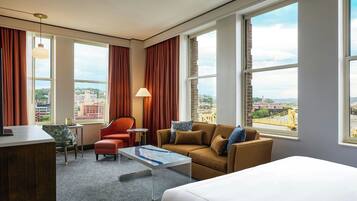 Quarto de hotel com cama, sofá, poltrona, mesa pequena e abajur. Janelas amplas com cortinas e vista para a cidade.
