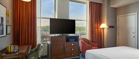 Quarto de hotel com cama, escrivaninha com cadeira, televisão e vista da cidade através de amplas janelas.