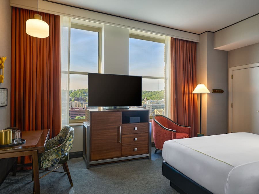 A hotel room with a bed, a desk with a chair, a television, and a view of the city through large windows.