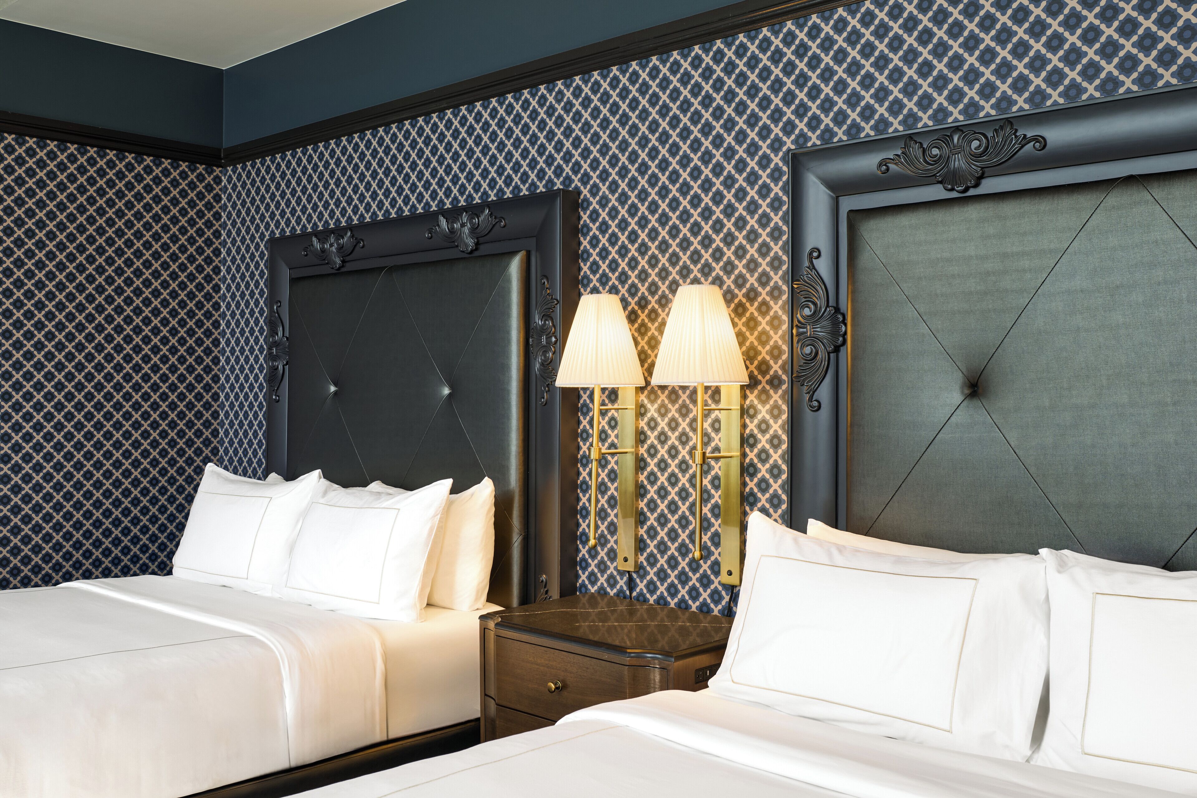 Quarto de hotel com duas camas, uma mesinha de cabeceira e uma luminária na parede.