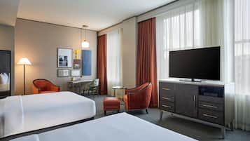 A hotel room with two beds, a desk, a chair, a television, and a large window with curtains.