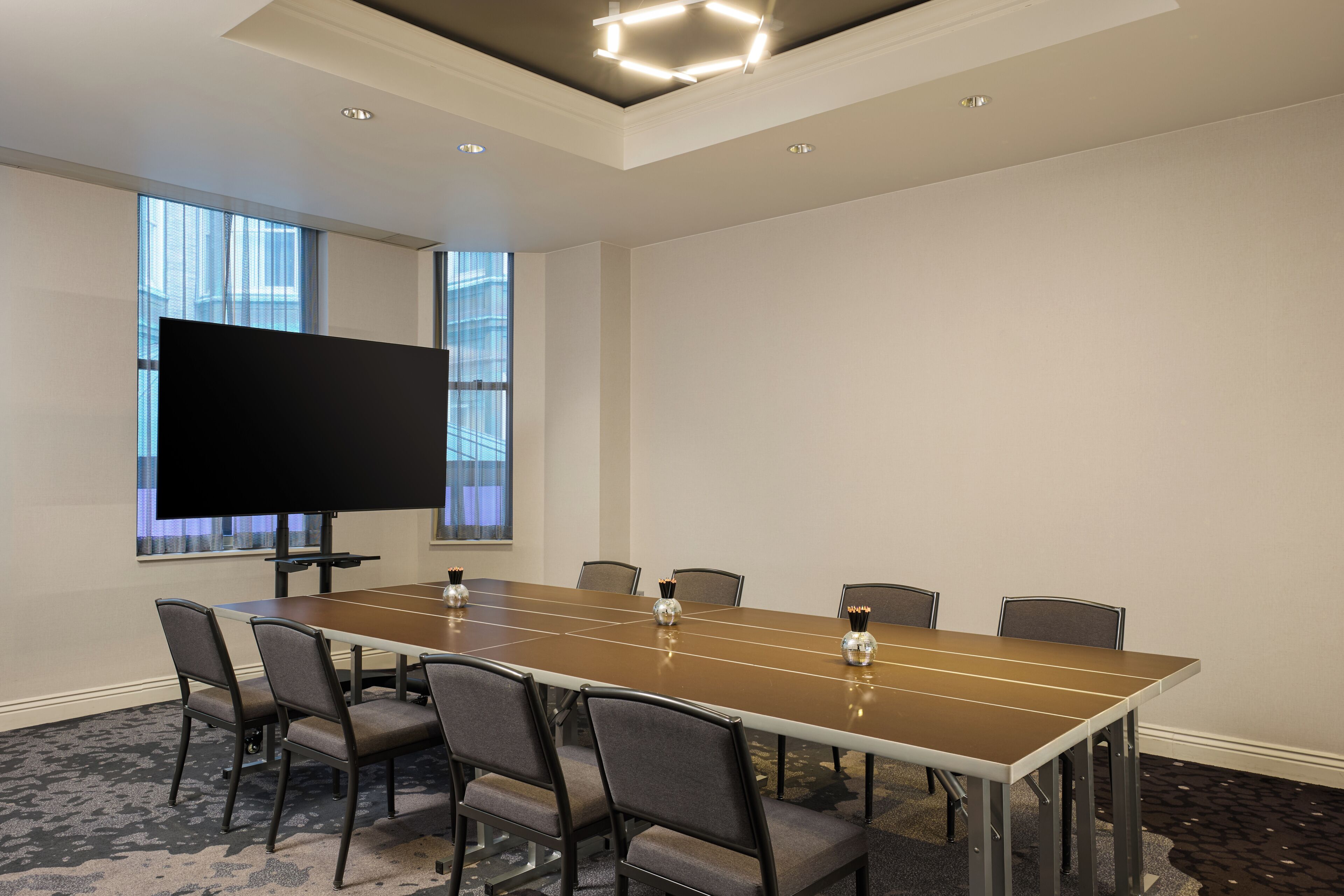 A conference room with a long table, six chairs, a flat-screen TV, and a window with blinds.