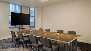 A conference room with a long table, six chairs, a flat-screen TV, and a window with blinds.