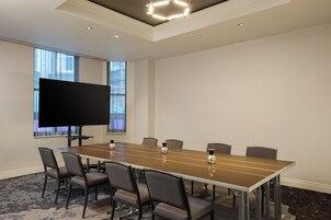 A conference room with a long table, six chairs, a flat-screen TV, and a window with blinds.