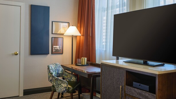 A hotel room with a television, a desk, a chair, a lamp, and a door.