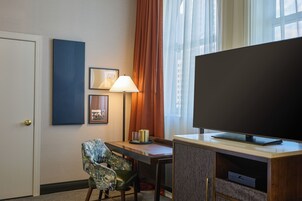 A hotel room with a television, a desk, a chair, a lamp, and a door.
