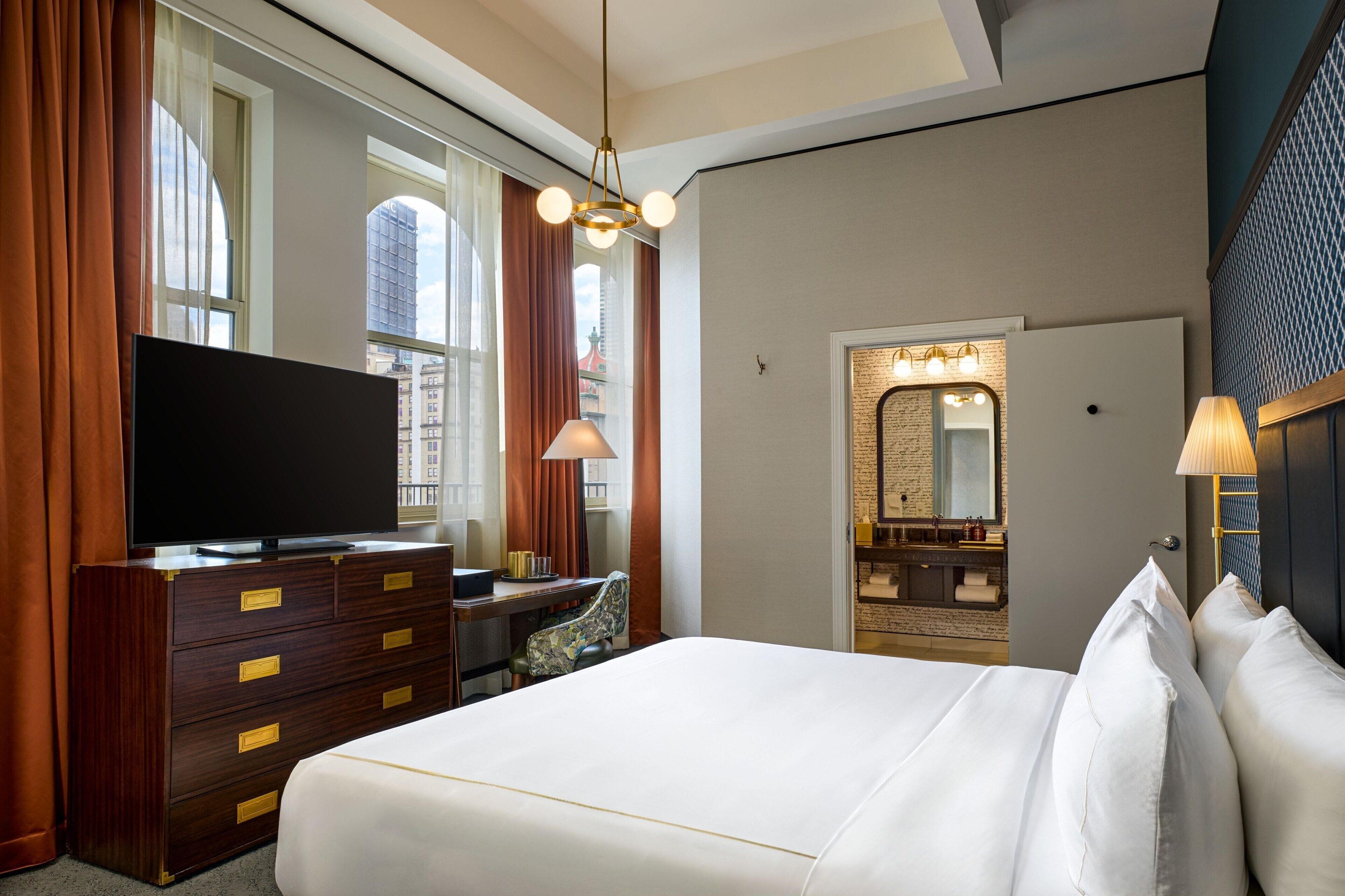 Une chambre d’hôtel dotée d’un grand lit, d’un téléviseur à écran plat, d’un bureau avec une chaise, d’une fenêtre avec des rideaux et d’une salle de bain avec un miroir et un lavabo.