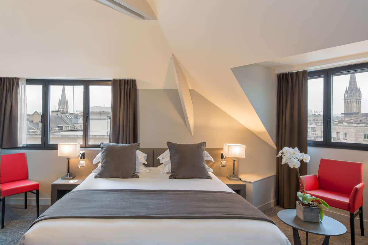 Une chambre d’hôtel avec un grand lit, deux tables de chevet, un fauteuil rouge, une petite table avec une plante et de grandes fenêtres donnant sur une cathédrale.