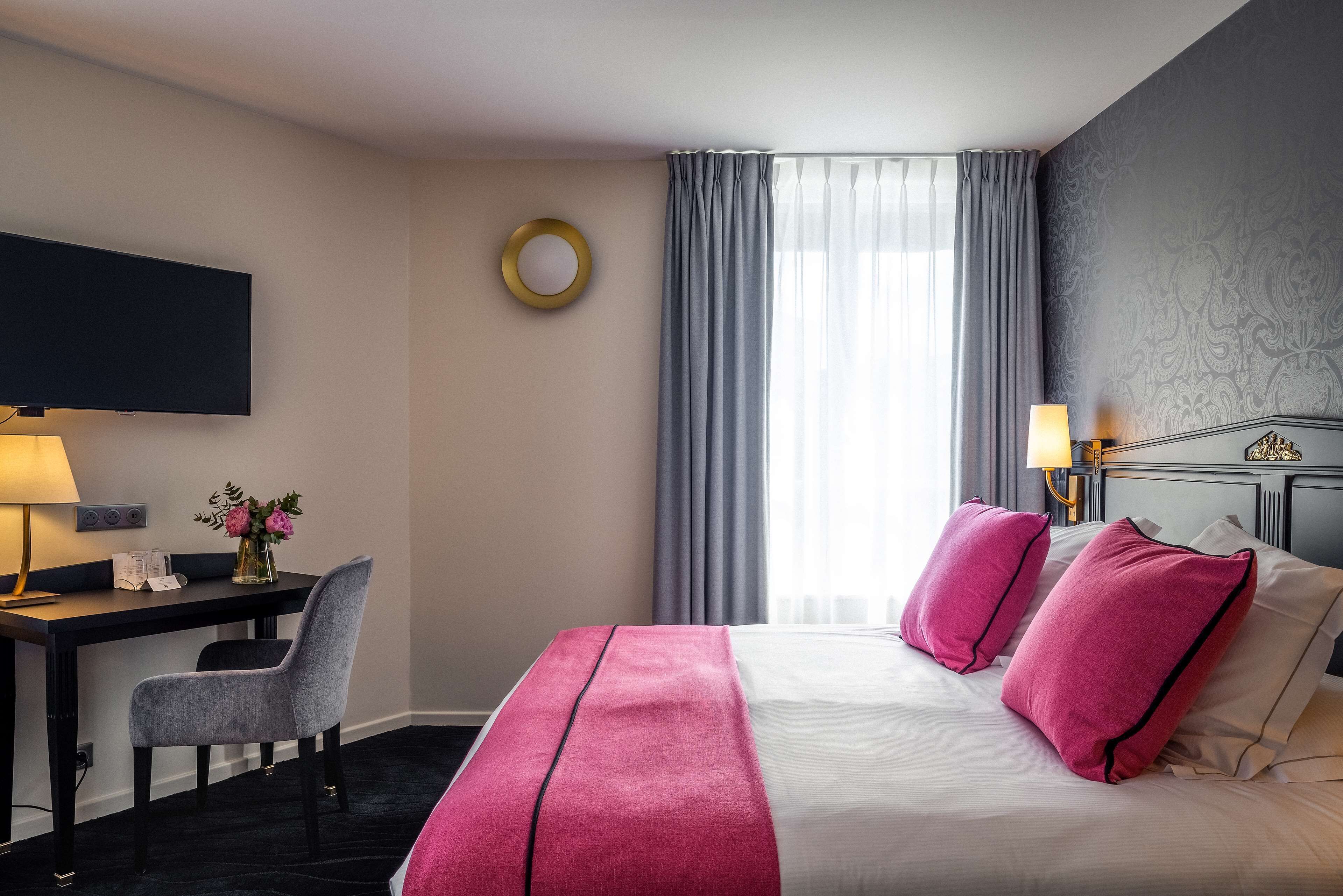 Quarto de hotel com cama, escrivaninha, cadeira, televisão e janela com cortinas.
