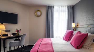 Quarto de hotel com cama, escrivaninha, cadeira, televisão e janela com cortinas.