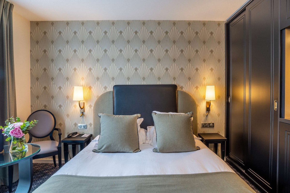 Une chambre d’hôtel avec un grand lit, deux lampes de chevet, une chaise, une petite table et un bureau avec un téléphone.