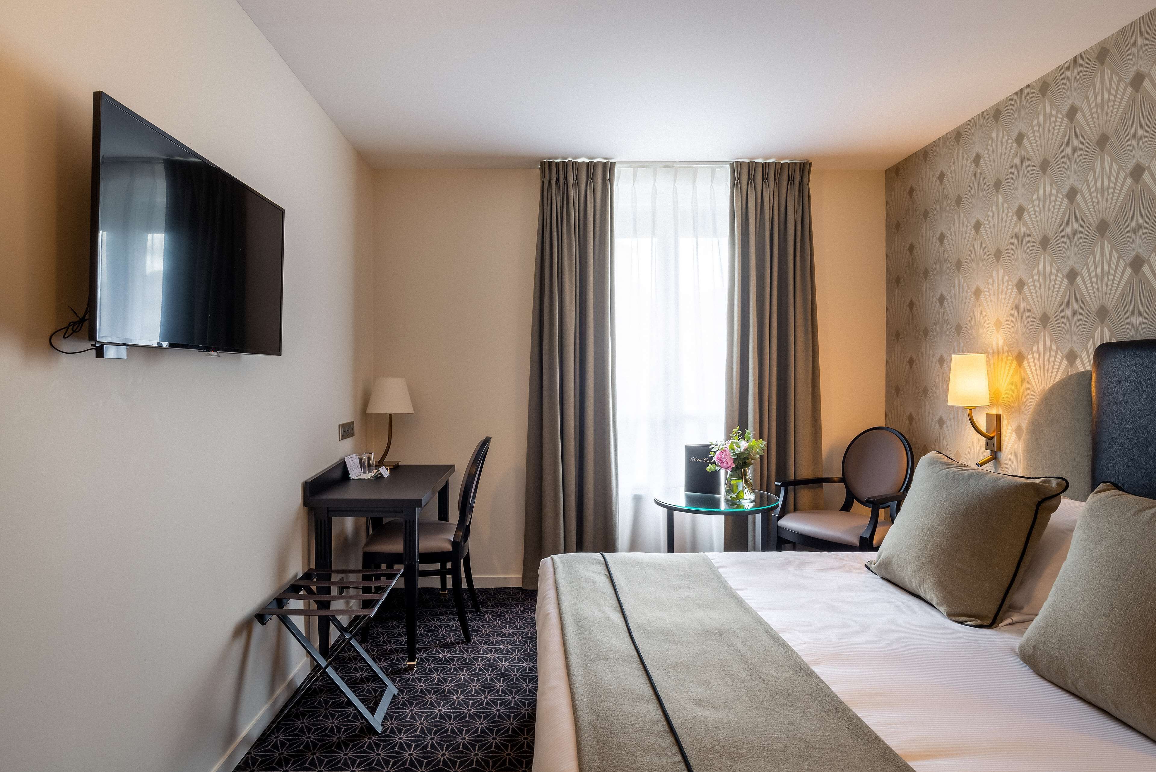 Ein Hotelzimmer mit einem Bett, einem Schreibtisch mit Stuhl, einem an der Wand montierten Fernseher, einem Fenster mit Vorhängen und einer Lampe auf dem Nachttisch.