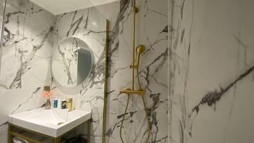 A bathroom with a marble wall, a round mirror, a gold showerhead, and a white sink with toiletries.