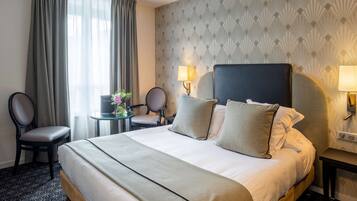 Una habitación de hotel con una cama grande, dos sillas, una mesita con un jarrón y una lámpara.