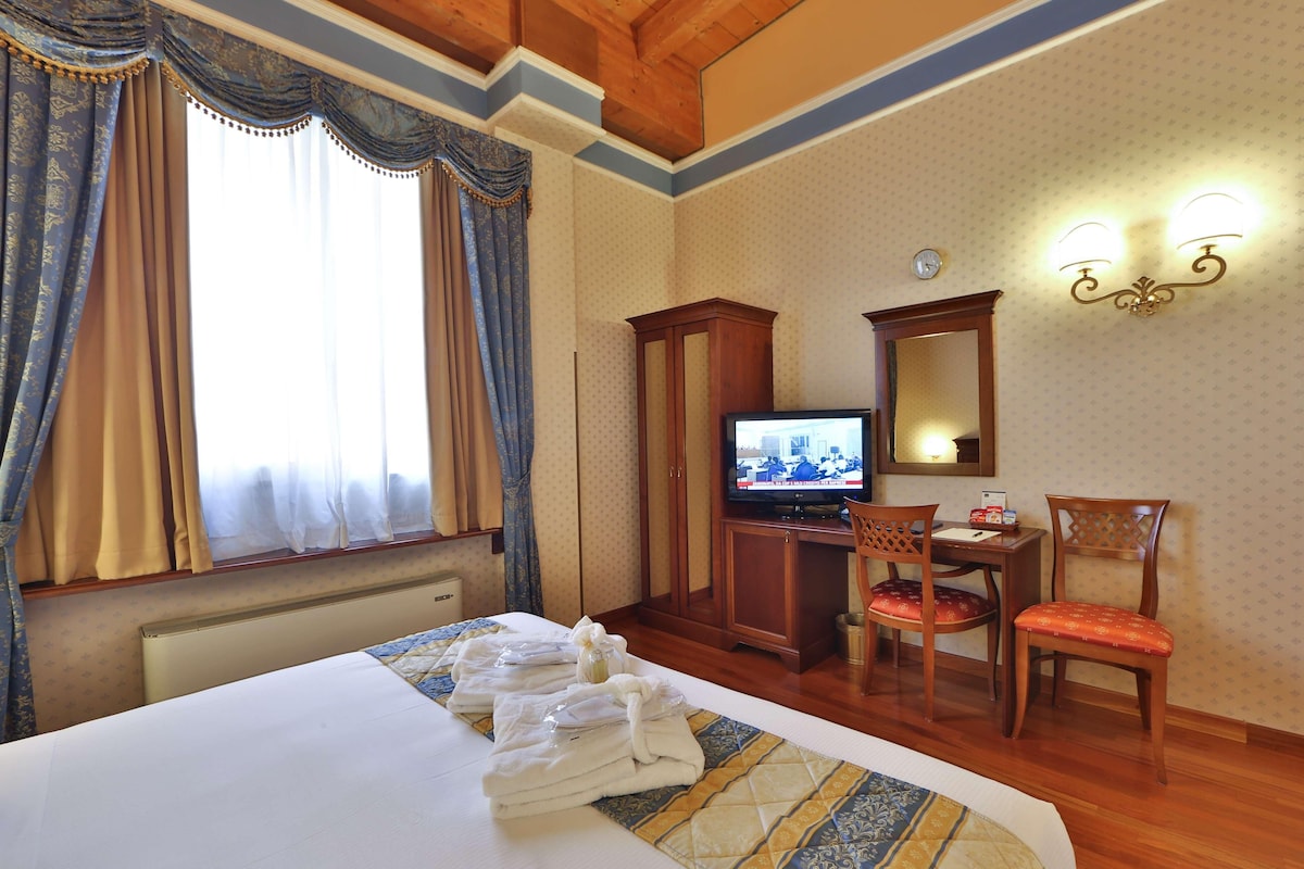 Habitació d'hotel amb llit, televisió, escriptori amb cadira i finestra amb cortines.