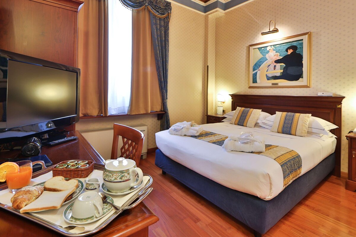 Een hotelkamer met een bed, een televisie, een bureau met ontbijtspullen en een schilderij aan de muur.