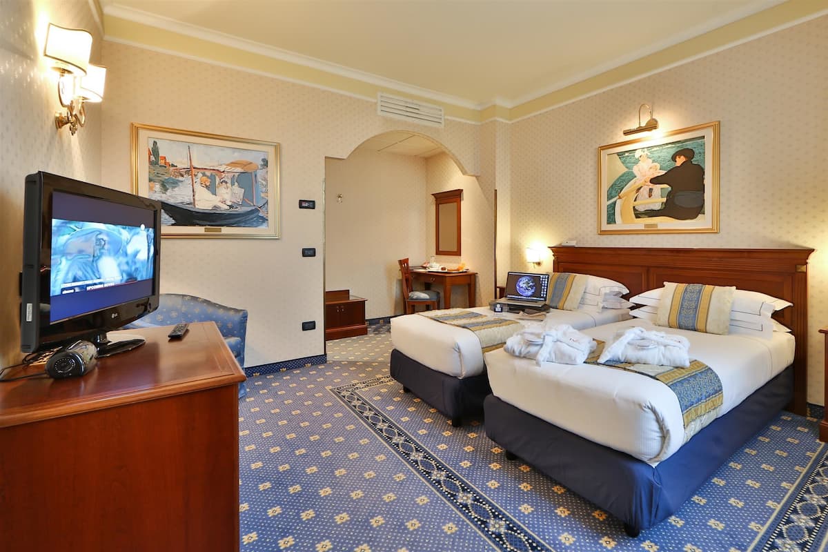 Habitación de hotel con dos camas, un escritorio con ordenador, televisor y obras de arte en las paredes.