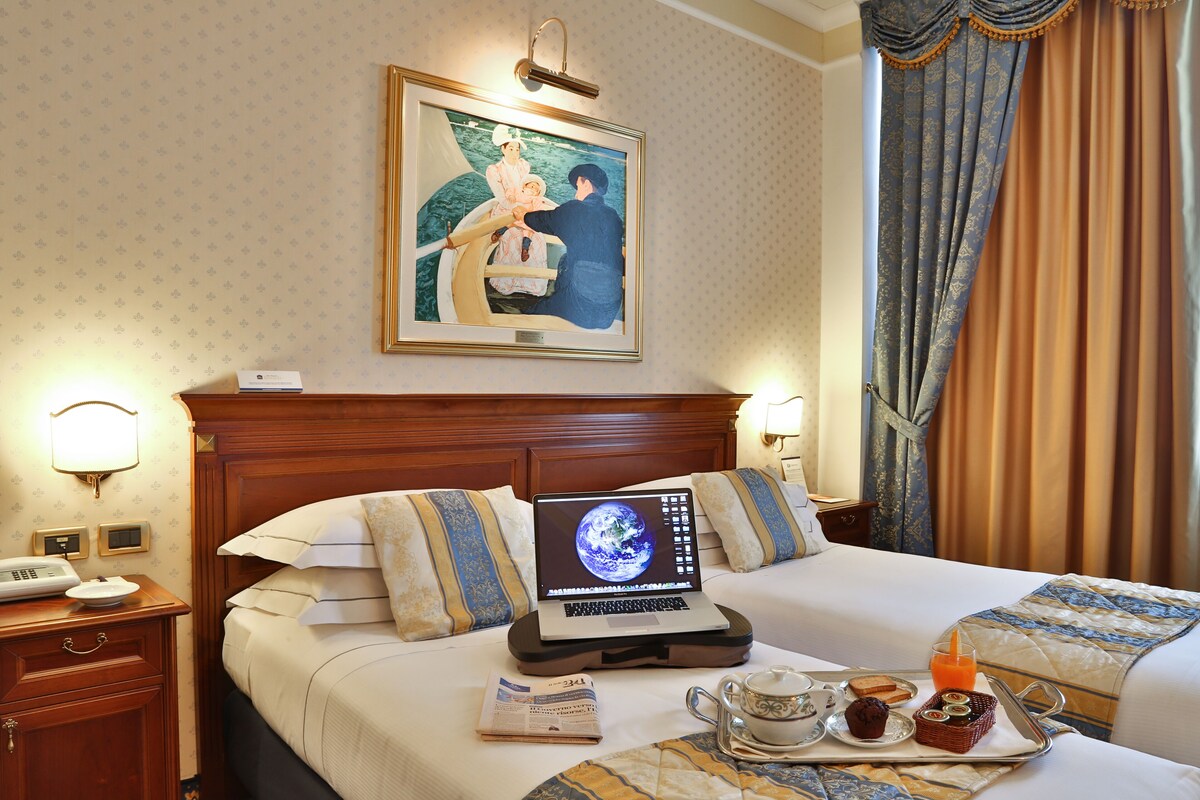 A hotel room with a bed, a laptop on a bedside table, a tray with breakfast items, and a painting on the wall.